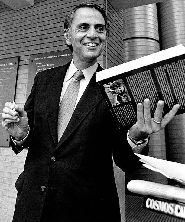 астробиолог Карл Саган с книгой в руках улыбается.