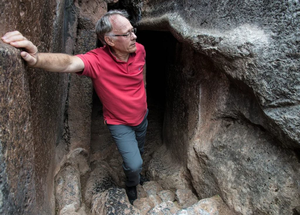 Писатель Грэм Хэнкок выходит из пещеры в красной рубашке.